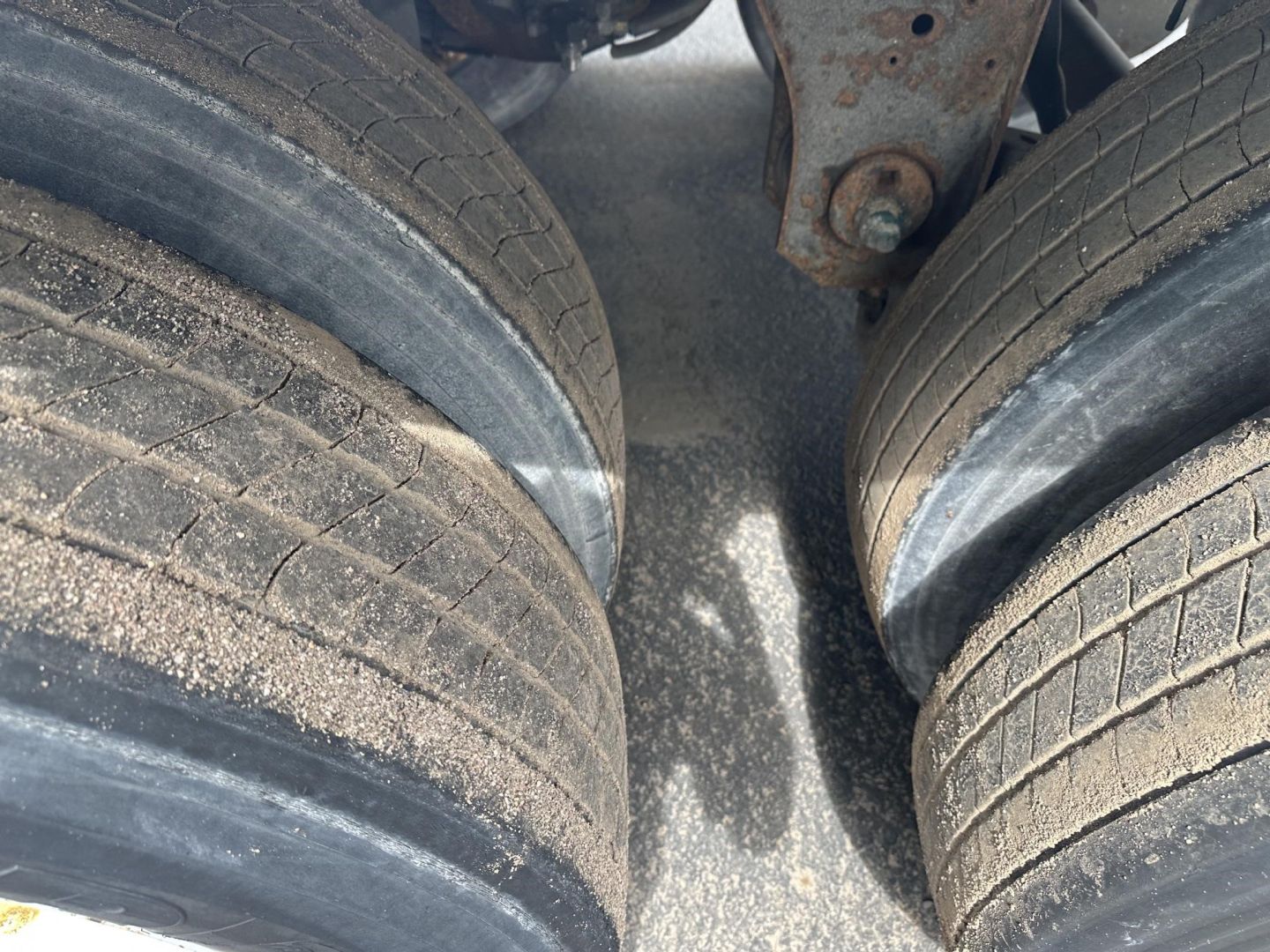 Dual rear tires of a utility trailer with gravel/dust on the road surface between tires; portion of frame and suspension components visible.