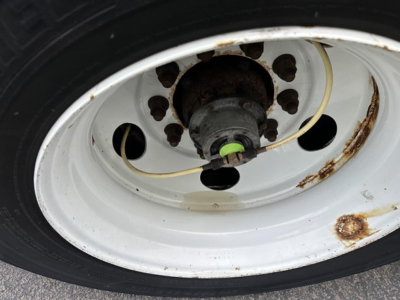 Trailer wheel area showing white steel rim with black tire, rusty hub area, and a green valve or cap with a light beige hose nearby.