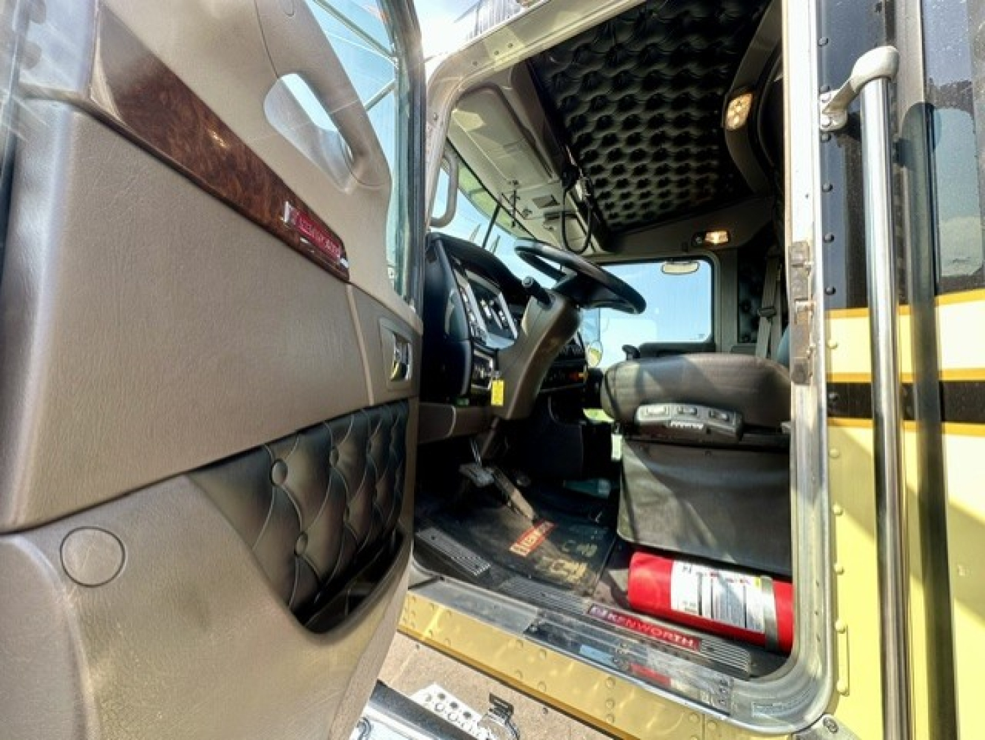 Kenworth W900L truck interior cabin view from open driver door showing steering wheel, dashboard, seat, and floor mat; engine bay not visible.