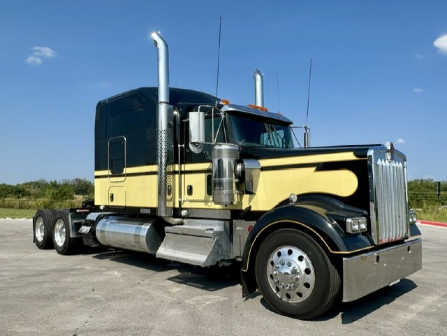 Kenworth W900L truck with black and gold paint, chrome grille, dual exhaust stacks, and polished fuel tanks.
