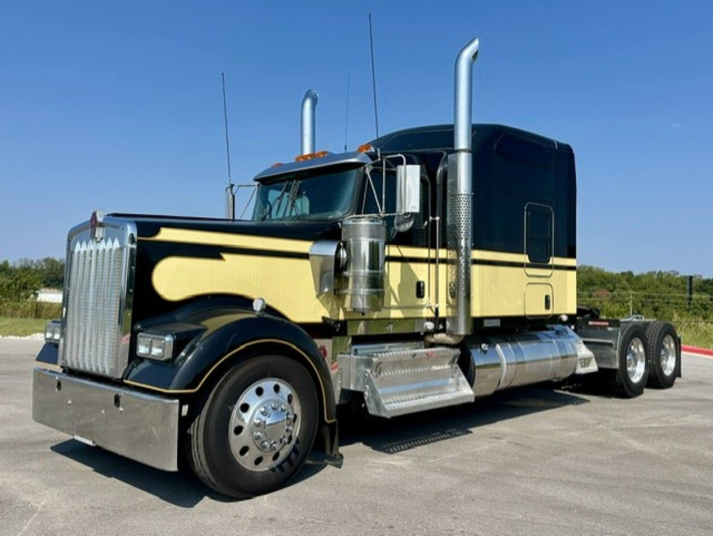 Kenworth W900L conventional sleeper truck in black and tan with chrome exhaust stacks, grille, and dual rear wheels. Side view showing cab with vertical exhausts and aluminum fuel tanks.