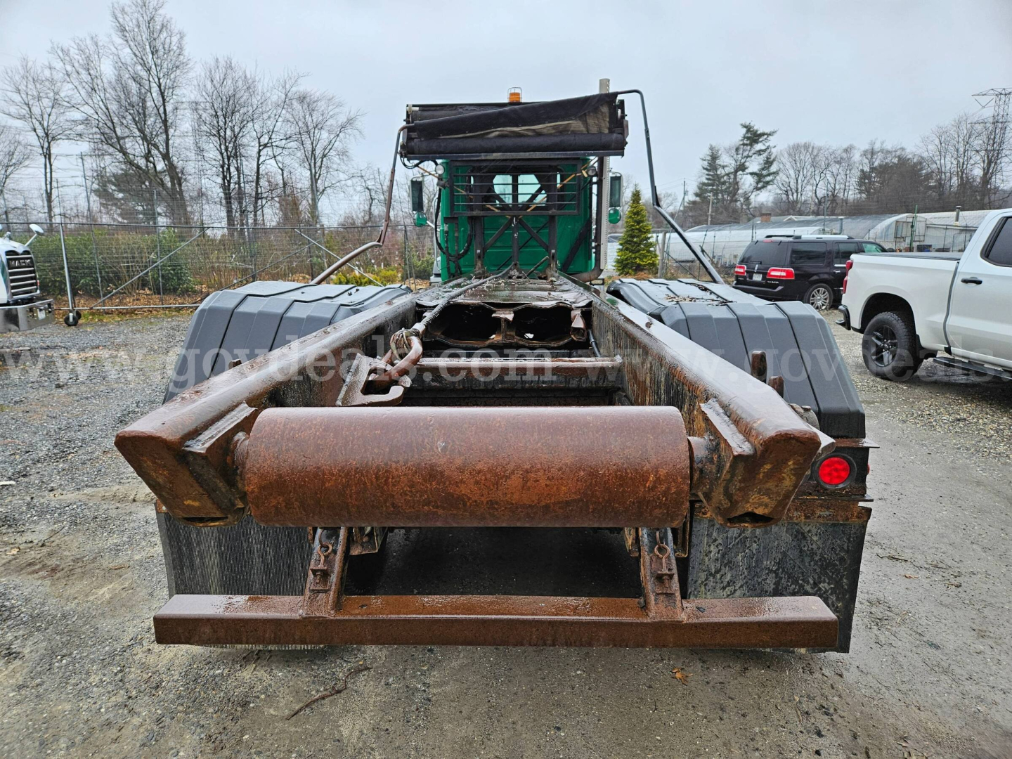 Rear view of a used 2008 Mack GU813 roll-off truck showing rusty hitch and frame components. Visible chain, hydraulic arms, and rear bumper with taillight on right.