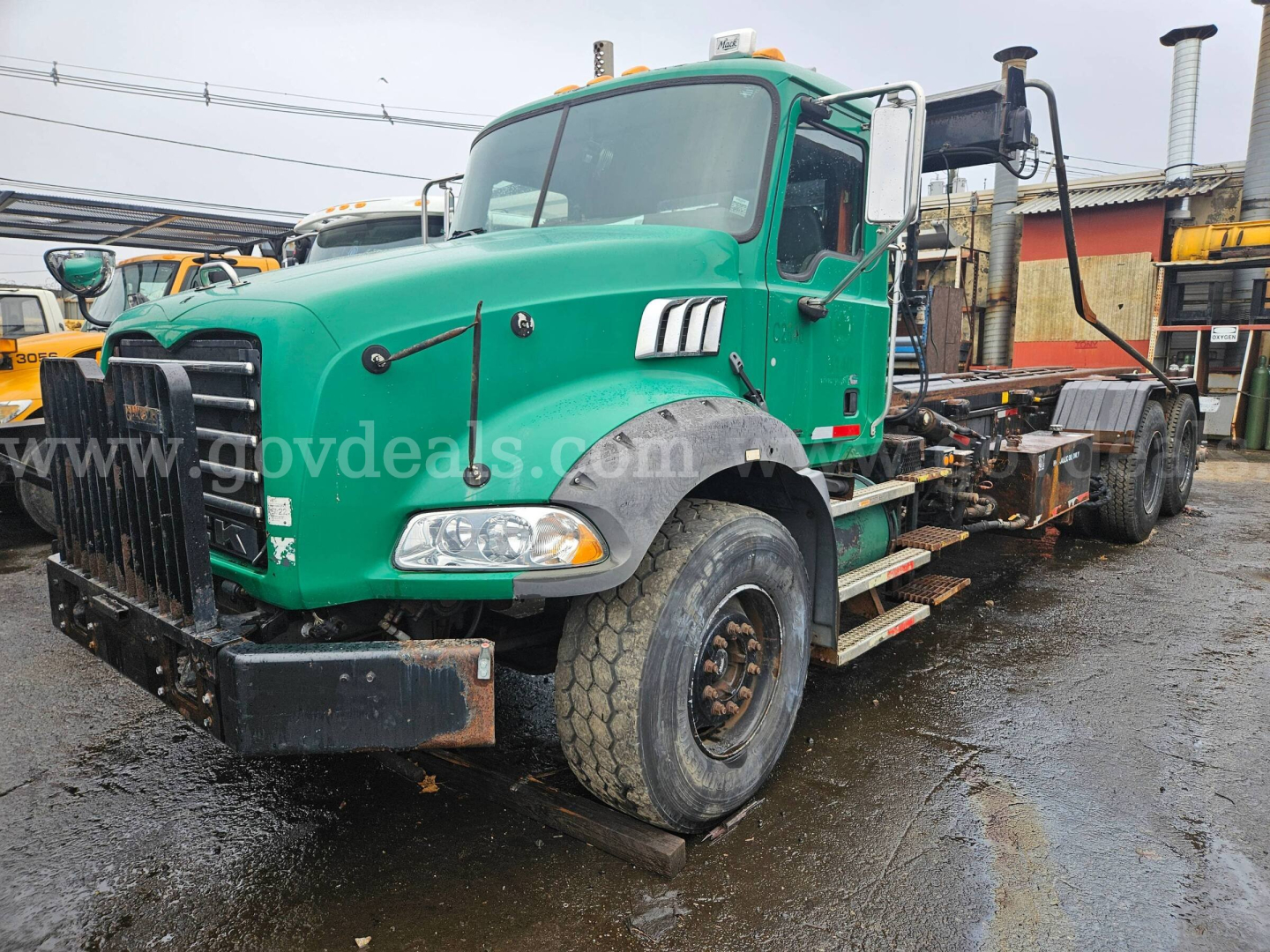 Green 2008 Mack GU813 roll-off truck showing front grille, cab, and chassis with heavy-duty bumper and dual exhaust stacks in yard.