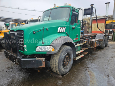 Green 2008 Mack GU813 roll-off truck showing front grille, cab, and chassis with heavy-duty bumper and dual exhaust stacks in yard.