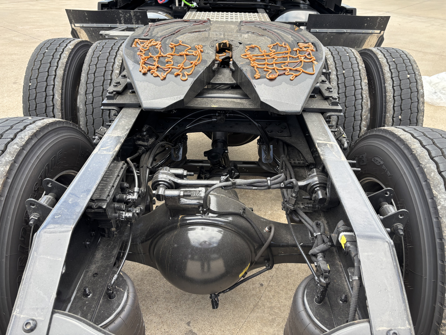 Rear view of a Volvo VNL truck chassis showing dual rear axles, suspension, and drive components. Visible text includes orange decorative markings on the fifth wheel guards and structural members, no readable model text.