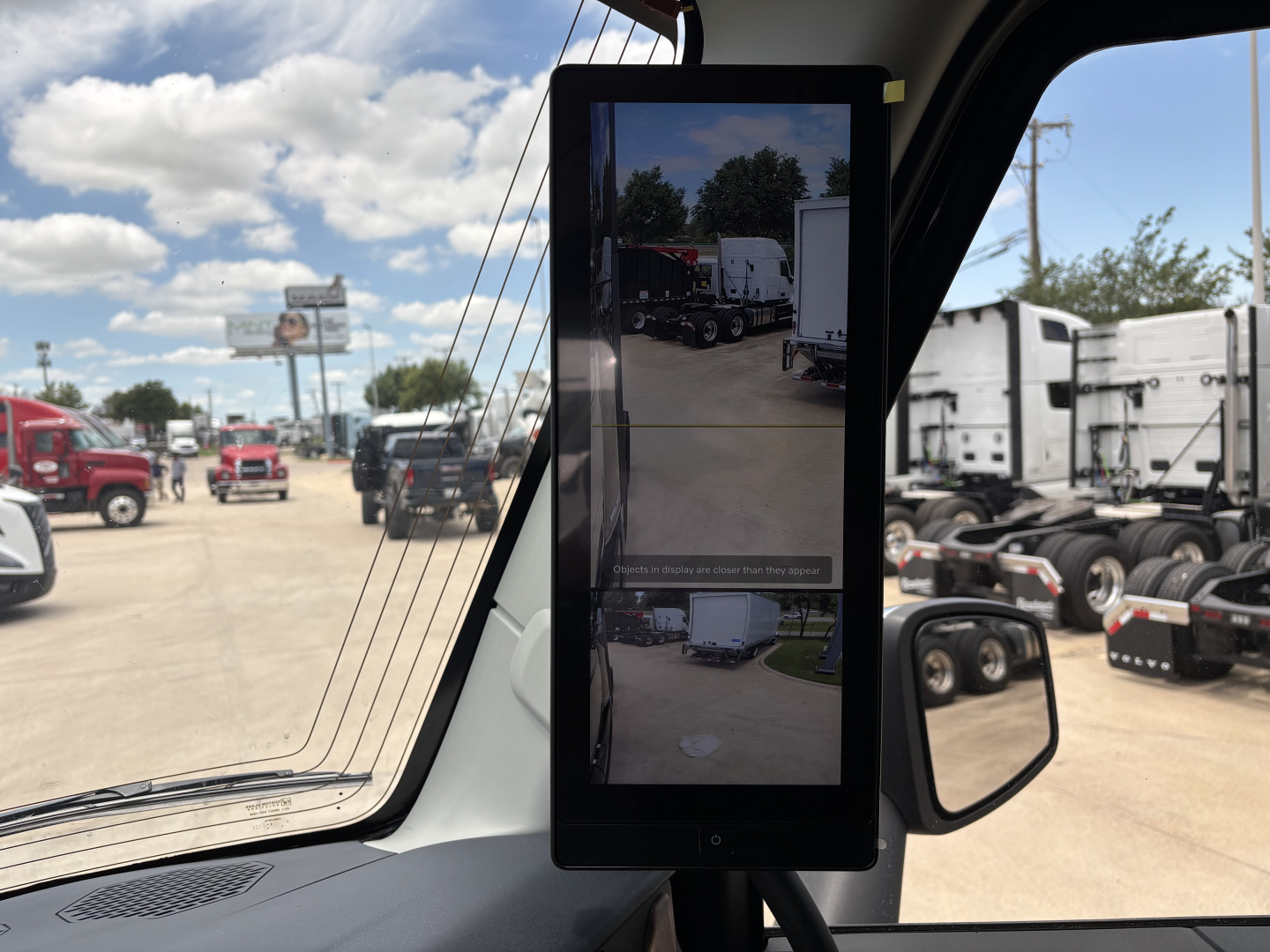 Truck cabin interior with a tall vertical tablet touchscreen device mounted near the windshield; outdoor lot with other trucks and trailers visible through the windshield and side window.