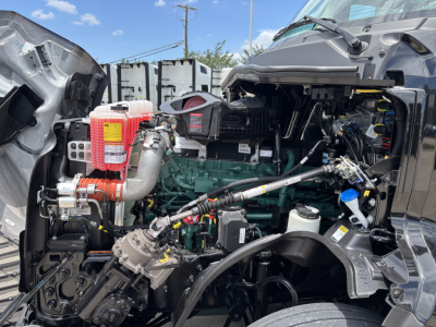 Vehicle engine bay of a Volvo VNL64T860 showing green engine, red coolant reservoir, air intake, hoses, and various hydraulic lines.
