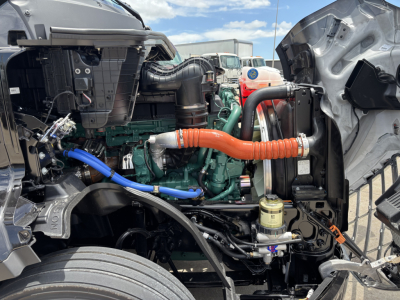 Volvo VNL64T860 engine bay visible with green engine block, orange intake hose, blue coolant hose, black radiator components, and various hoses plus yellow filter; wake of cab shows open hood area.