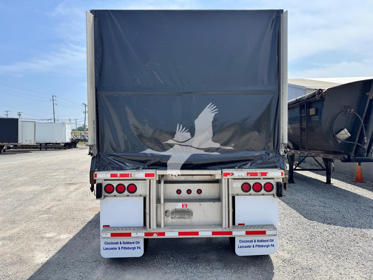 Trailer rear view showing aluminum flatbed curtainside with black tarp, taillights, and bumper with reflectors; logo bird decal on tarp; surrounding trucks in lot.