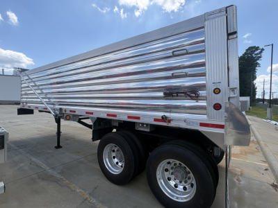 End dump trailer with triple-axle chrome wheels, polished aluminum side panels, and rear tailgate. Side has tie-down bars and reflective striping; VIN badge and stock number not visible.