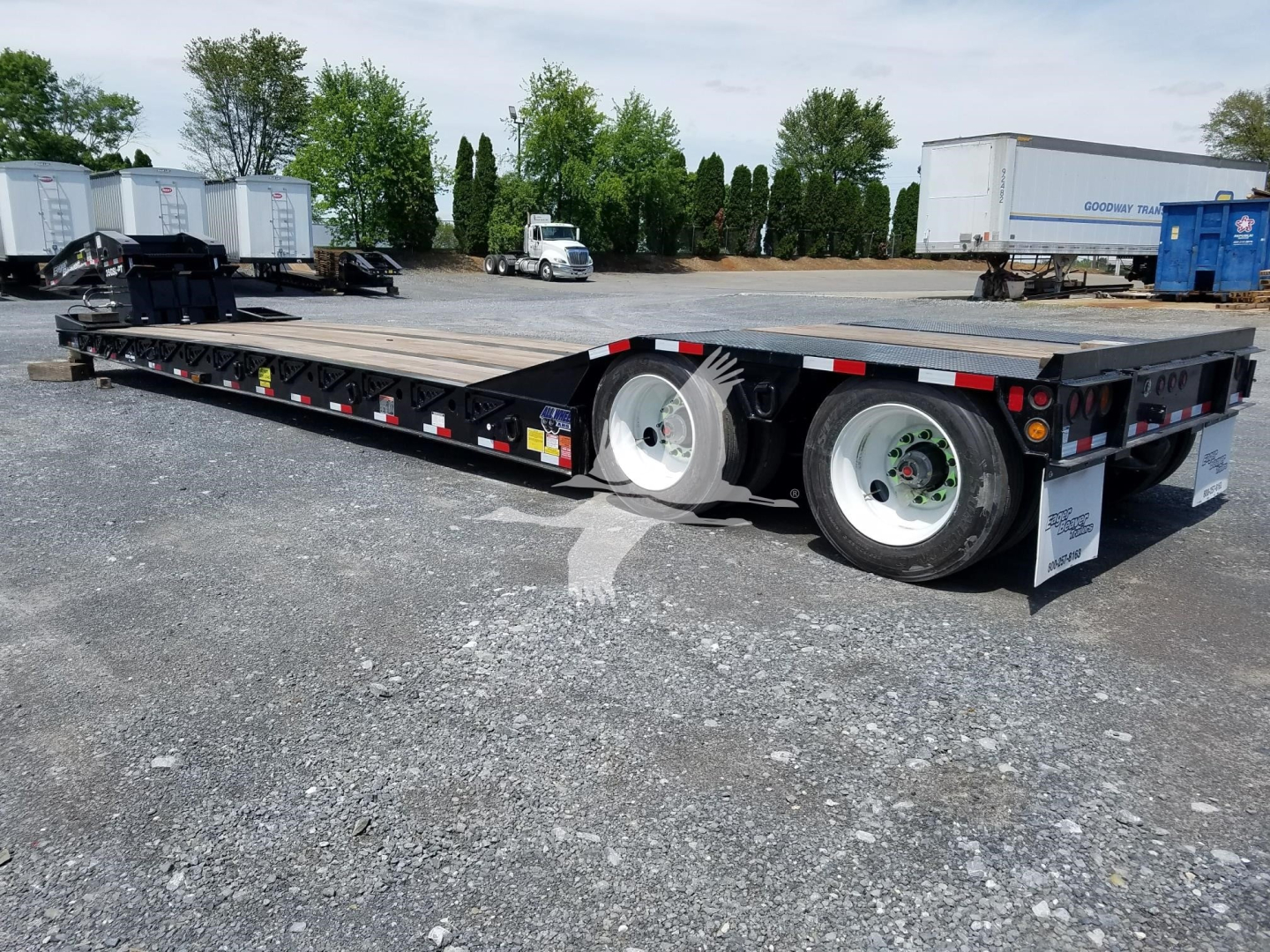 Trailer: lowboy trailer with dual rear axles and flatbed deck, showing white rims, black frame, reflective safety decals, and a Gooseneck hitch area.