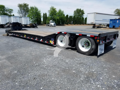 Trailer: lowboy trailer with dual rear axles and flatbed deck, showing white rims, black frame, reflective safety decals, and a Gooseneck hitch area.