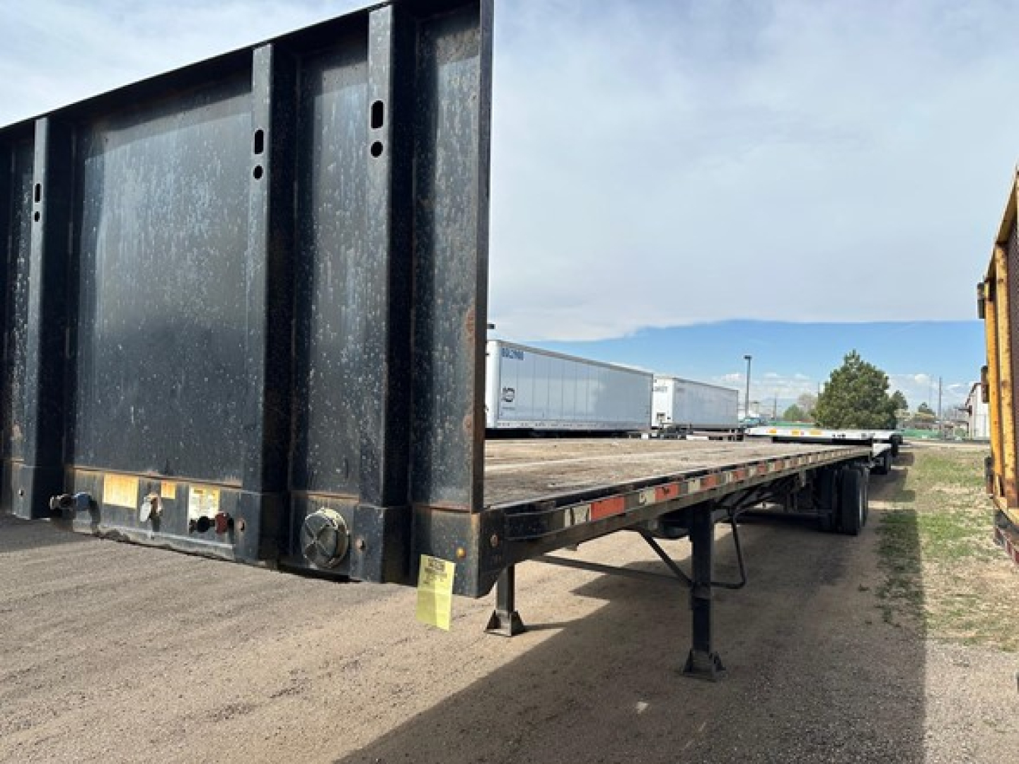 Flatbed trailer side and rear showing hydraulic ramp/door mechanism and support legs with black steel frame and yellow tags; Clark steel flatbed trailer, used condition.