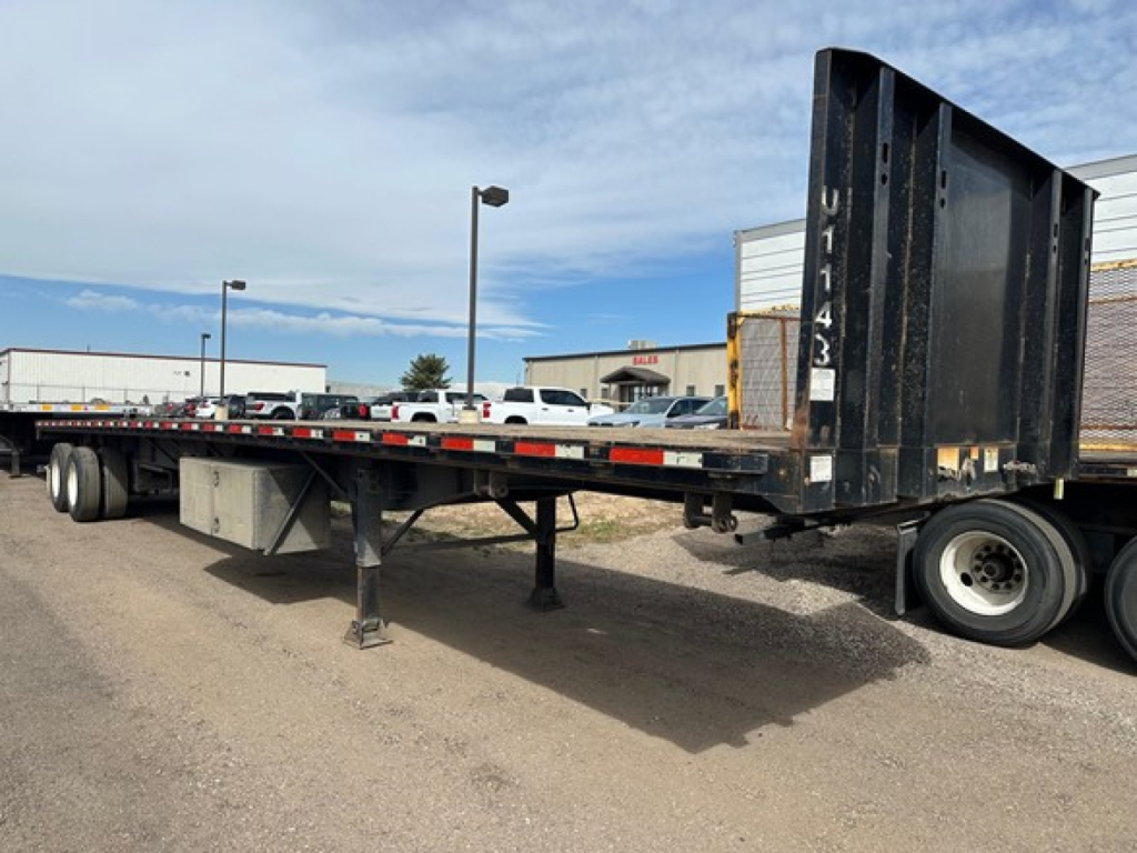 Flatbed trailer showing gooseneck-style rear with tall ramp, side rails, and landing gear in a parking lot.