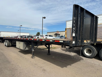 Flatbed trailer showing gooseneck-style rear with tall ramp, side rails, and landing gear in a parking lot.