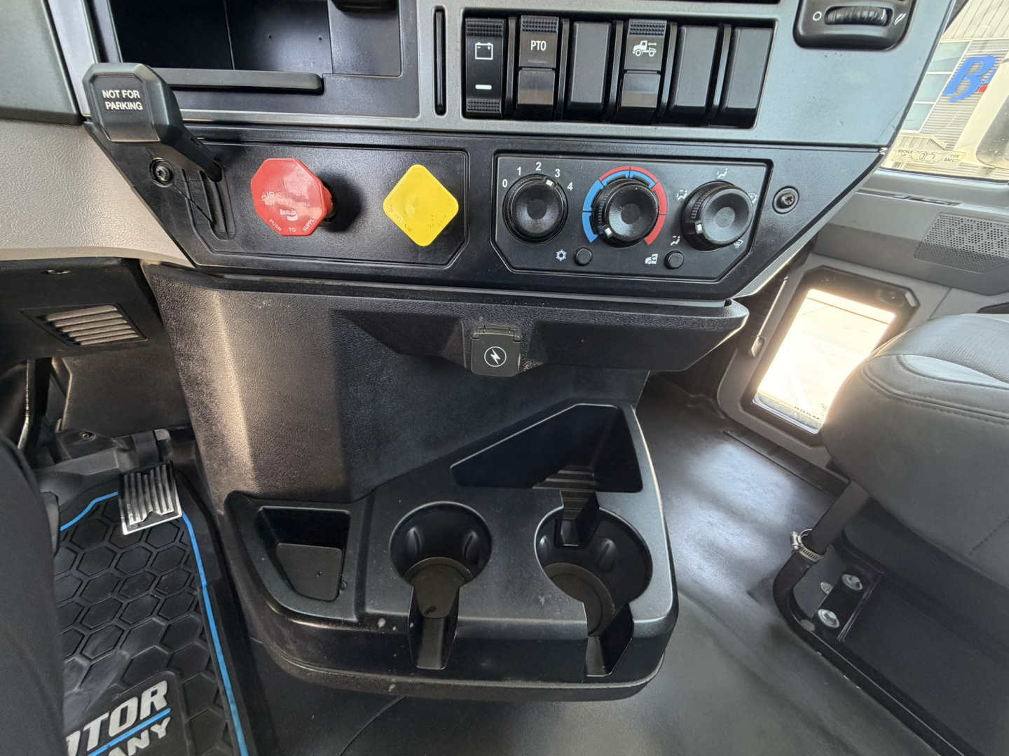 Dashboard area of a Mack PI64T showing control dials, switches, cup holders, and pedal area. Visible text includes 'NOT FOR HARM' on red knob, and circular climate controls.