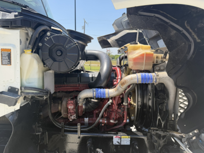Mack PI64T engine bay showing powertrain and coolant reservoir with hoses and belts visible.