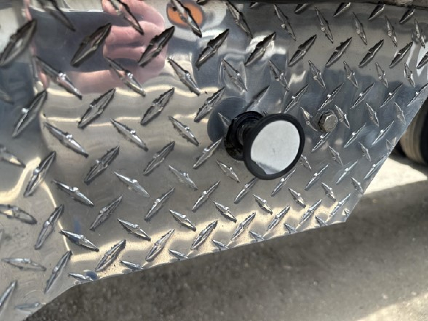 Diamond plate trailer side with a circular black-and-silver plug or cap protruding from the metal surface, chrome finish reflecting light.
