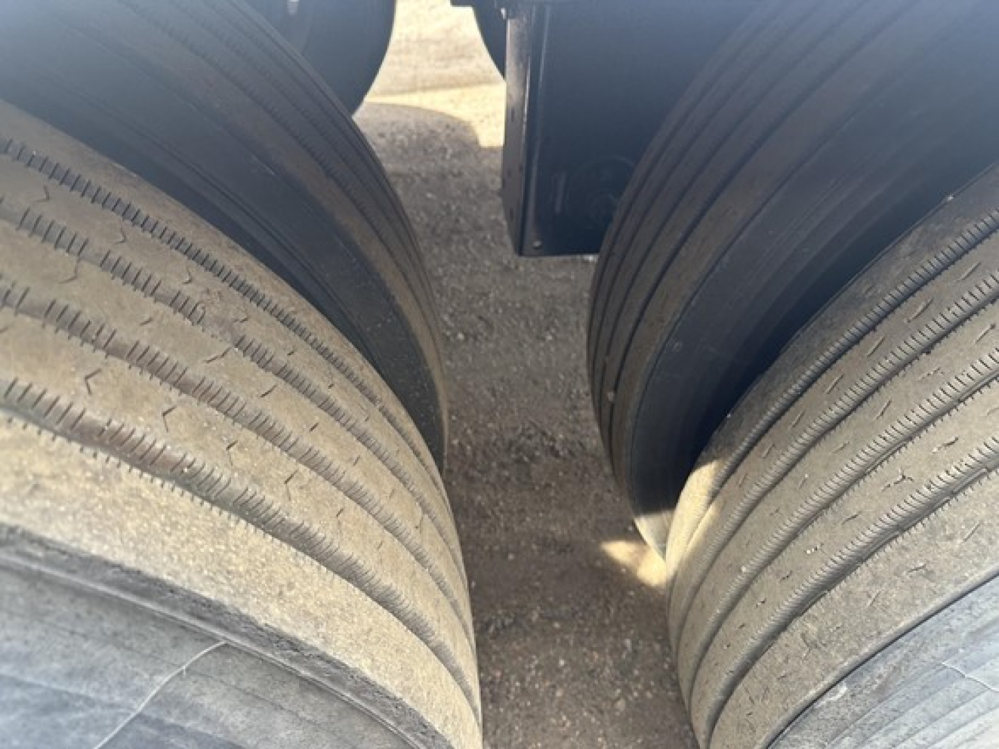Dual rear tires of a dump trailer with visible treads and dirt along the sidewalls, central gap between tires, undercarriage shadow visible.