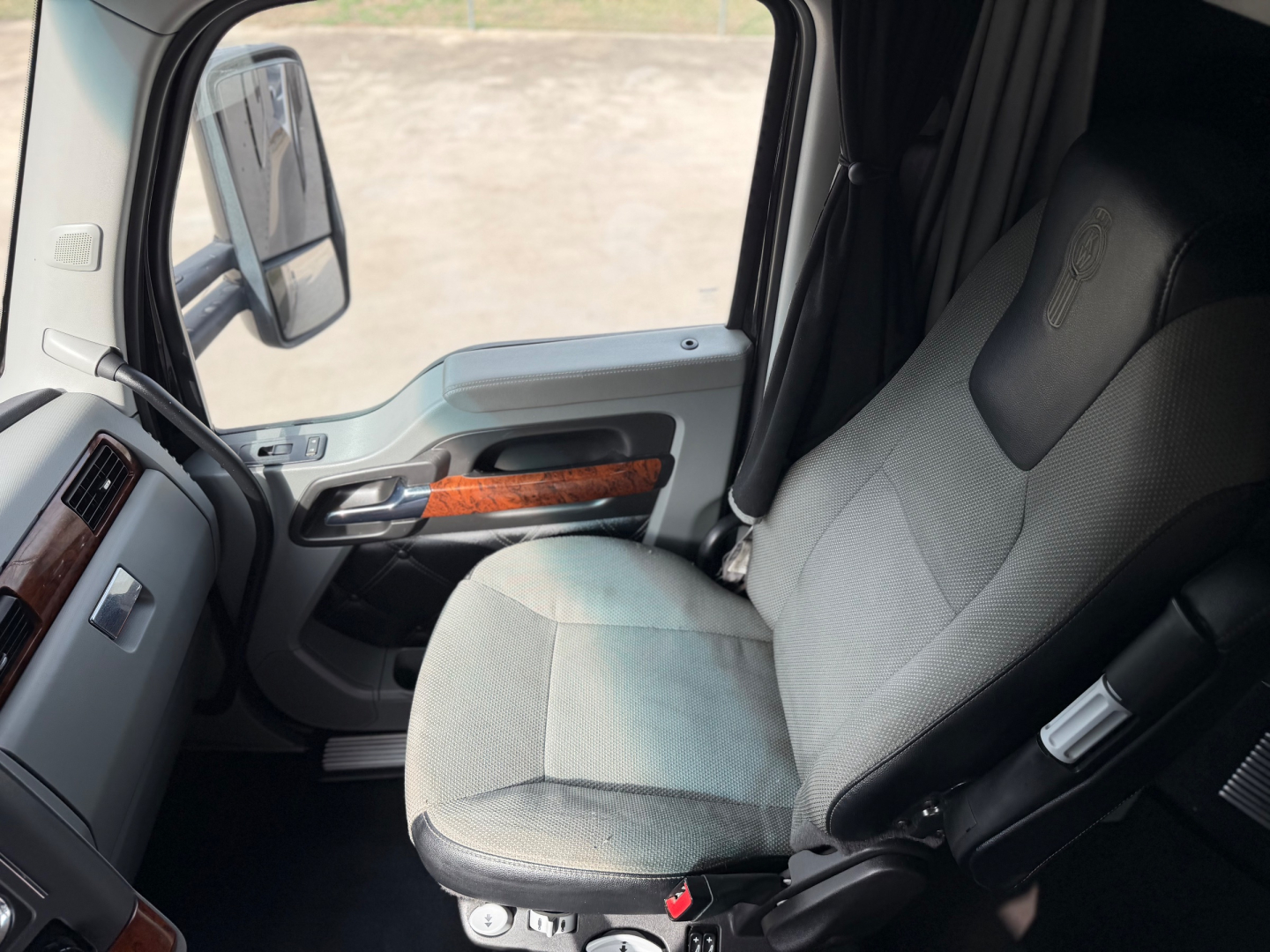 Kenworth T680 truck cabin interior with gray fabric seat, steering area, and door panel showing wood trim and controls