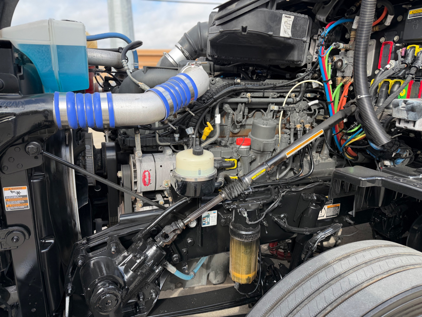 Kenworth T680 engine bay showing air intake tube, wiring harness, hydraulic reservoir, fuel filter, and various hoses and components.