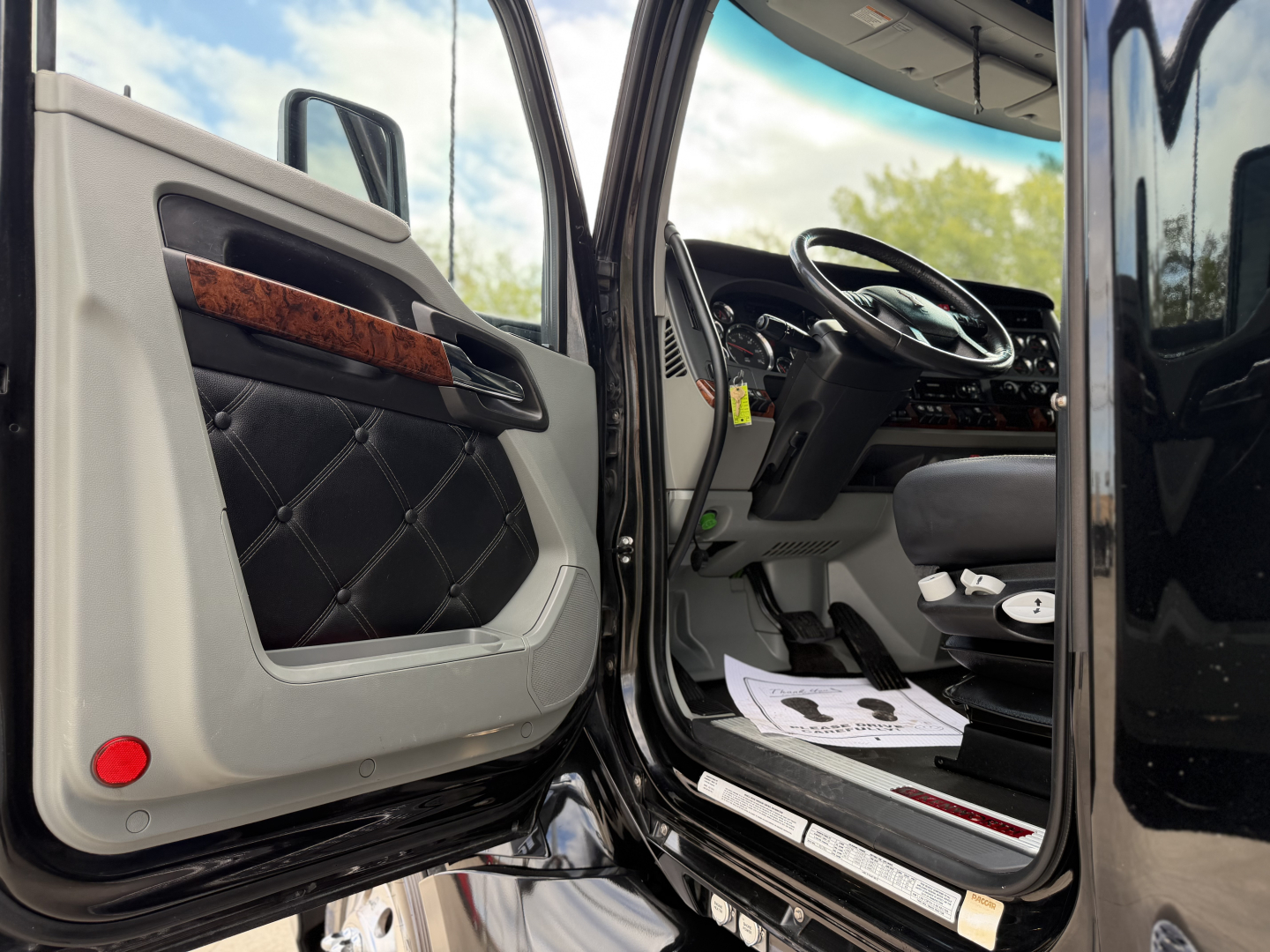 Truck cab interior door open of Kenworth T680 showing driver seat and steering wheel with door panel featuring black quilted insert and wood trim
