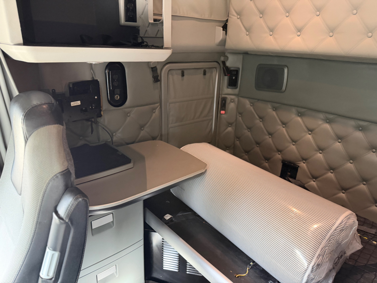 Kenworth T680 sleeper cab interior showing quilted side panels, bench-style bunk, table, and control panel with small monitor.