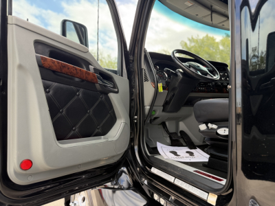 Truck cab interior door open of Kenworth T680 showing driver seat and steering wheel with door panel featuring black quilted insert and wood trim