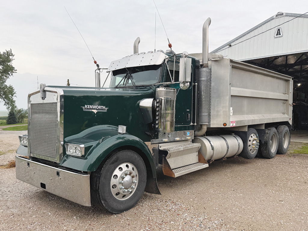 Kenworth W900L dump truck in green with chrome bumper, side tank, and large dual rear axles; Kenworth emblem on hood; dump bed visible.