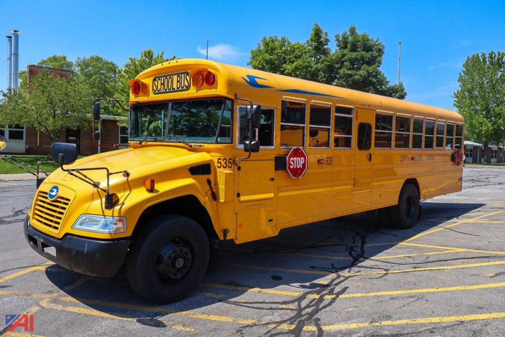Yellow school bus with Stop sign and blue bird logo on hood, front left view, roof signage and doors visible