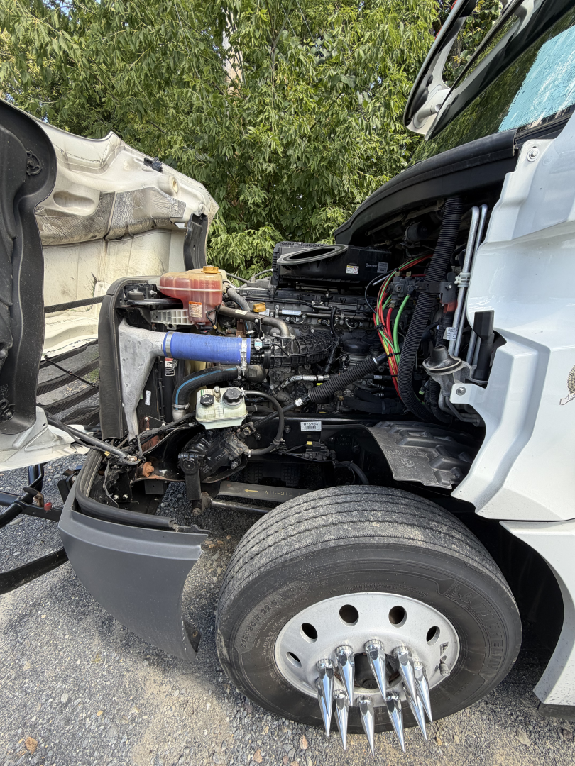 Truck engine bay of a Freightliner Cascadia with exposed diesel engine and front wheel; visible components include blue coolant hose, wiring harness, air intake area, and exposed engine bay accessories.