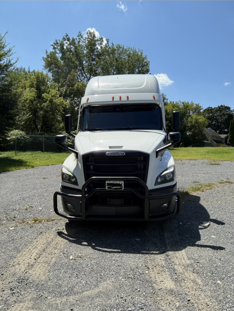 Truck front view showing Freightliner Cascadia with black grille, white cab, large bumper guard, side mirrors, and high cab. No visible text legible on the image.