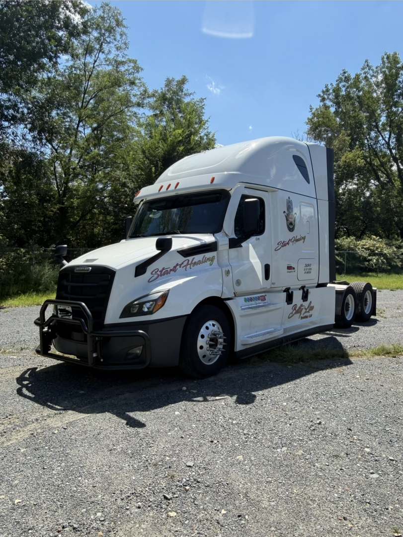 Freightliner Cascadia tractor-trailer in white with sleeper cab, front grill guard, and decals on doors and side.