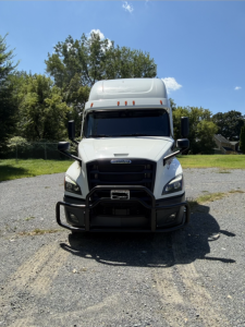 Truck front view showing Freightliner Cascadia with black grille, white cab, large bumper guard, side mirrors, and high cab. No visible text legible on the image.