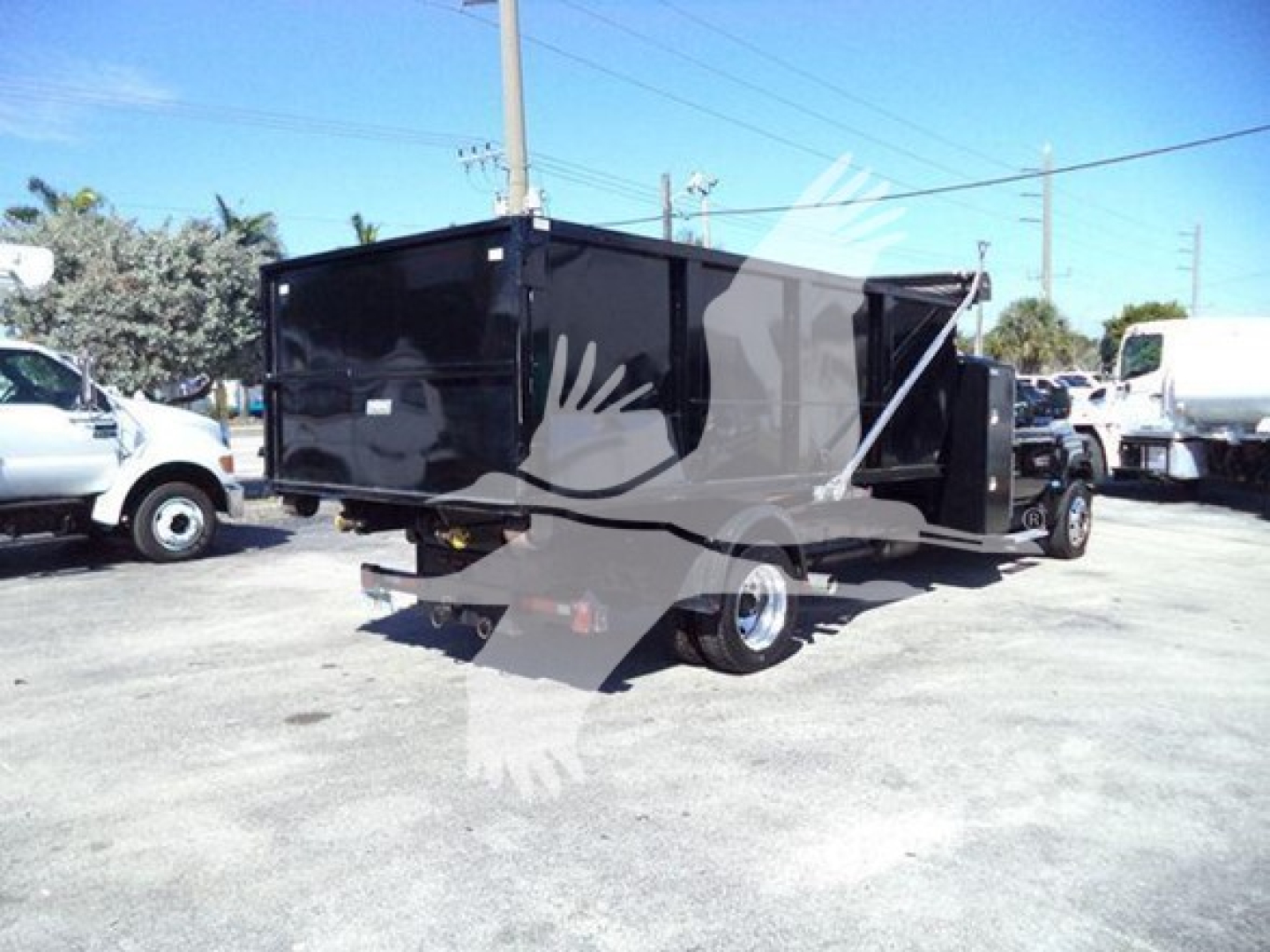 Truck: 2023 Chevrolet Silverado 6500HD roll-off body in an outdoor lot, black dump-style container, rear view showing dual wheels and hitch.