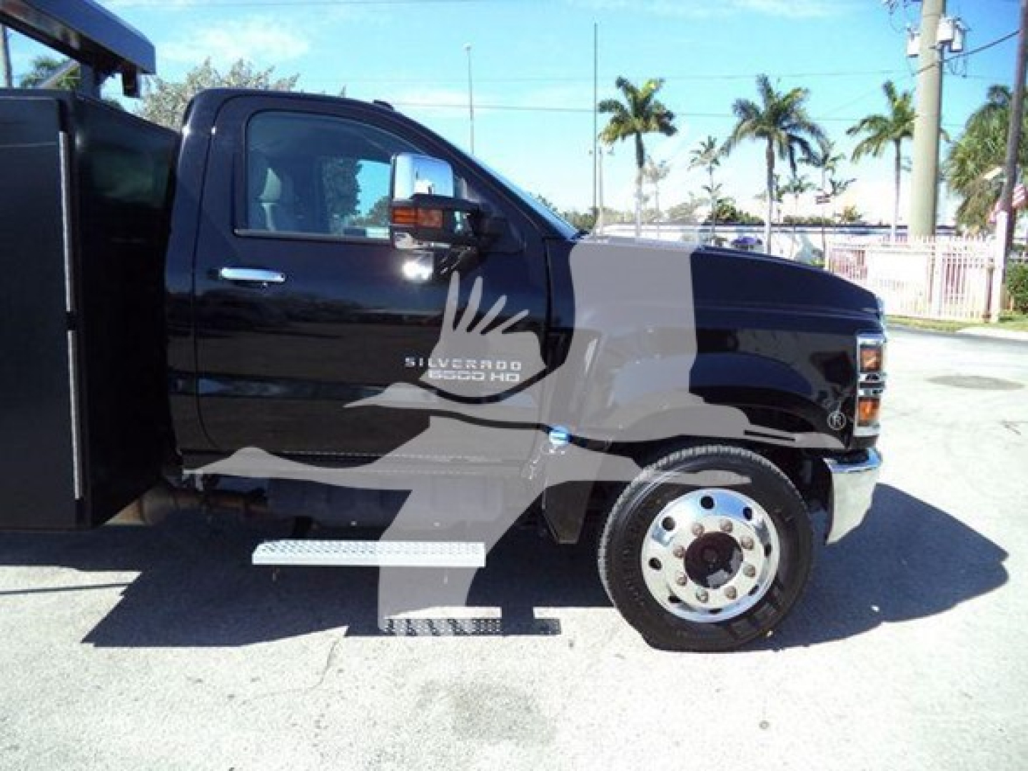 Truck side view showing 2023 Chevrolet Silverado 6500 HD logo on the door and chrome steps, black cab and front wheel, roll-off body in background.