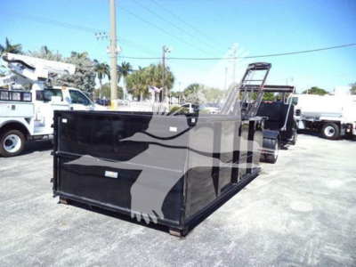 Truck bed container in a yard showing a black roll-off dumpster with white graphic, side view, with other equipment in background.