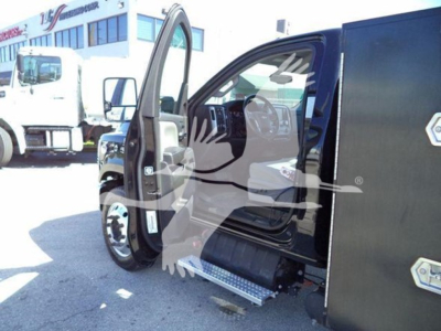 Truck cab interior with open door showing step rail and dashboard; portion of a black utility box on the side of a heavy-duty truck in a dealership lot.