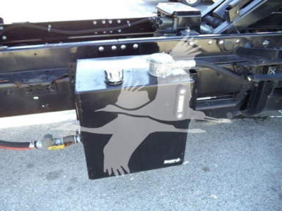 Aluminum/steel chassis with a rectangular black component mounted beneath the frame, electrical conduit and a hose visible, on a truck trailer chassis.