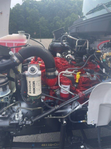 Hino L6 engine bay showing large red diesel engine, coolant reservoir, filters, hoses, and hydraulic lines.