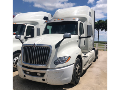 Truck cab and sleeper showing large chrome grille, headlights, side mirrors, and white exterior of an International LT625 tractor with two other trucks in background.
