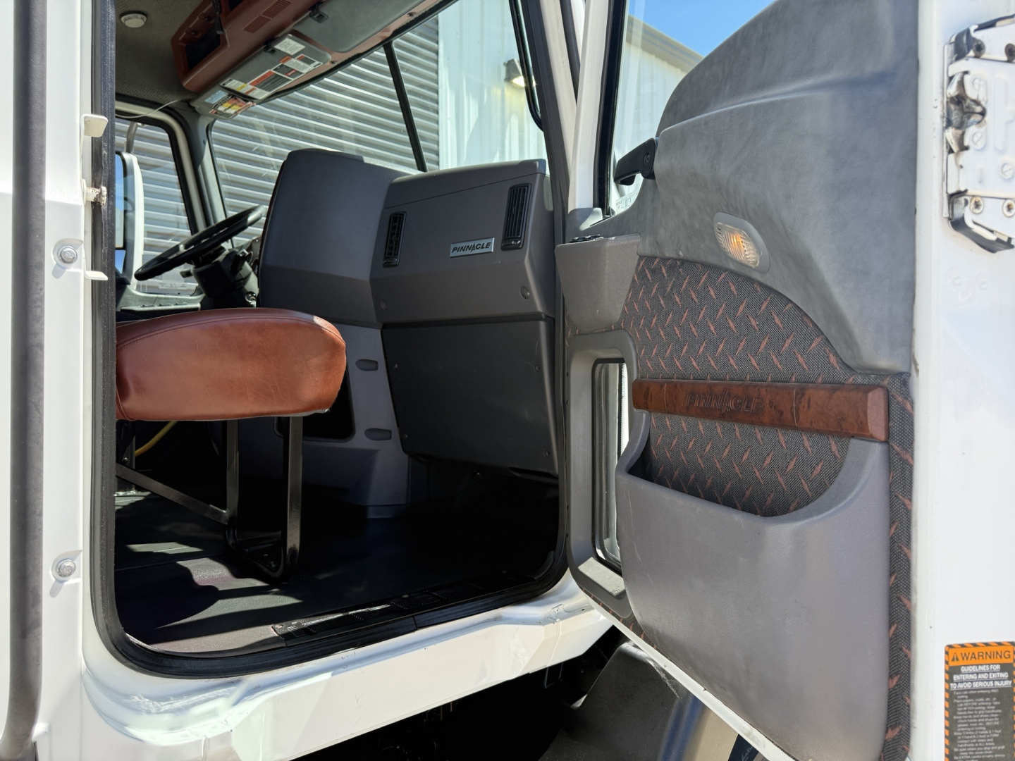 Cab interior of a 2018 Mack CXU612 daycab showing driver seat, steering wheel, dashboard, and open door with diamond plate panel.
