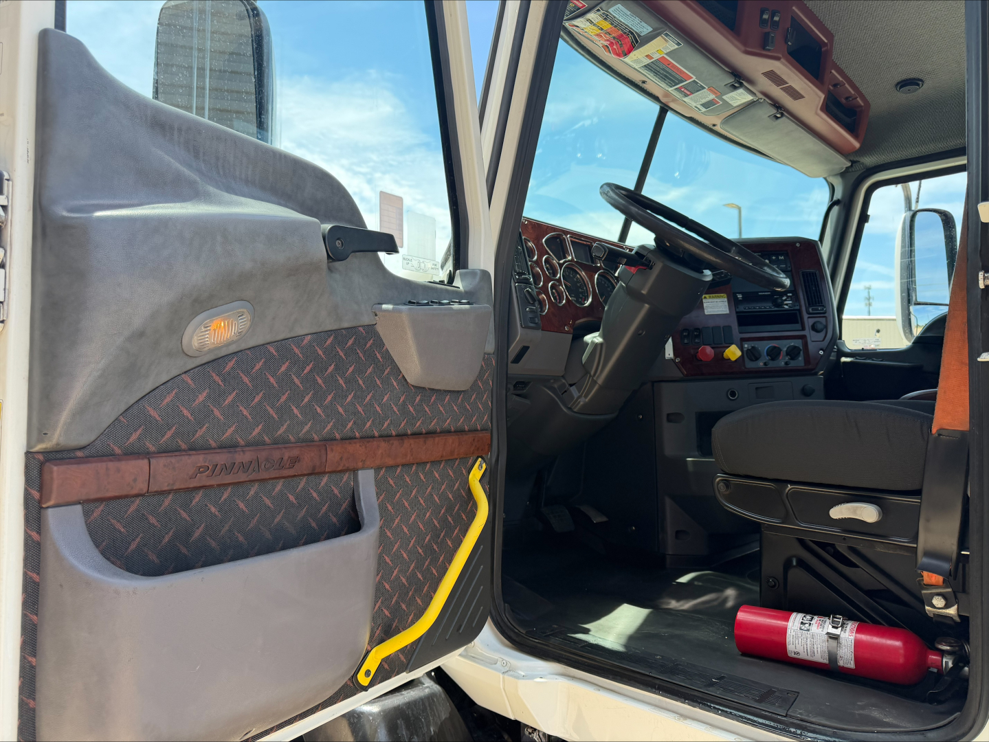 Truck cab interior with open door; dashboard, steering wheel, and driver seat visible. A fire extinguisher lies on the floor. Yellow handle on door panel. Diamond plate pattern on door panel.
