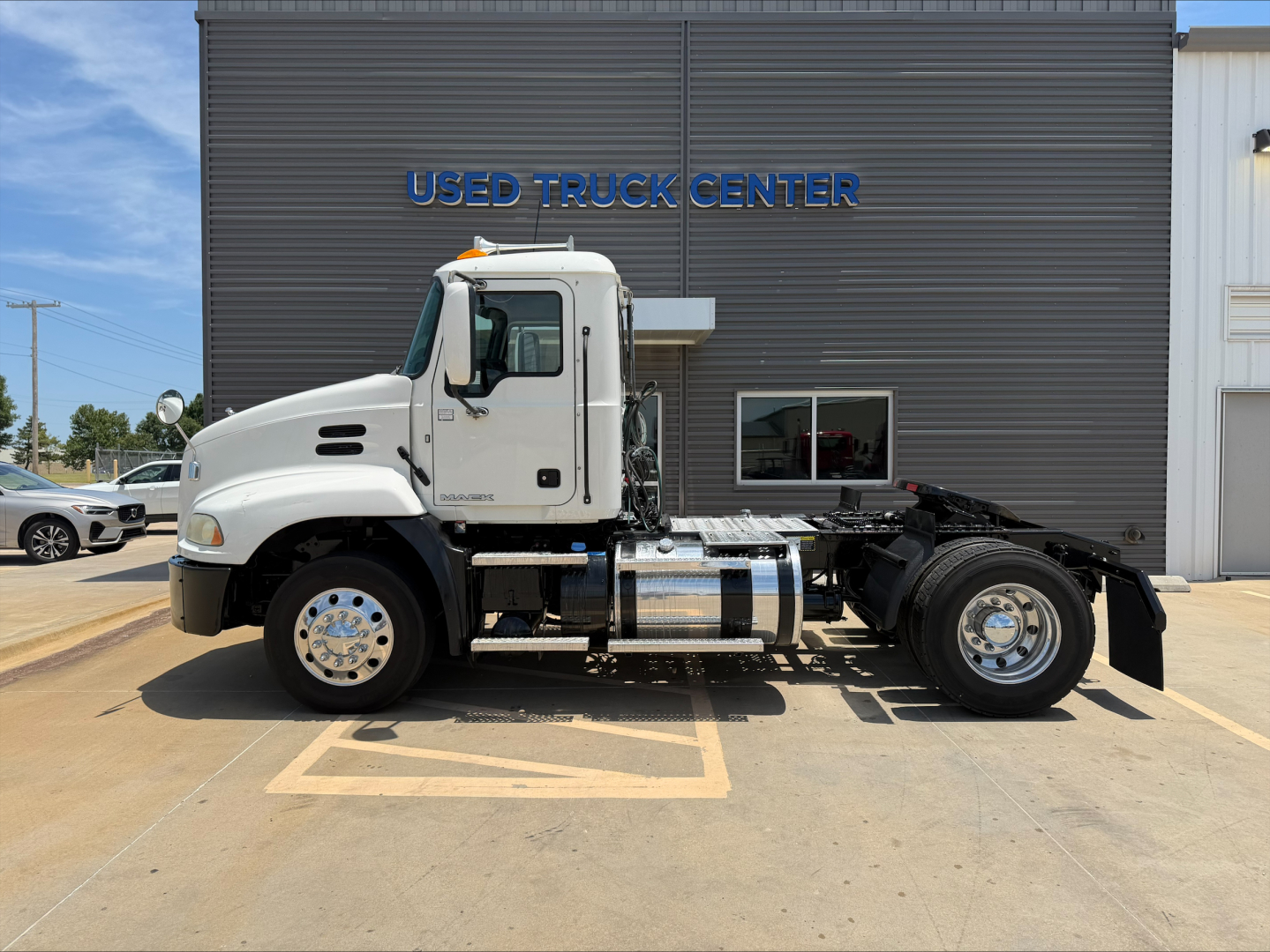 2018 Mack CXU612 conventional daycab truck side profile showing cab, chassis, fuel tanks, and wheels in front of a building with USED TRUCK CENTER signage.