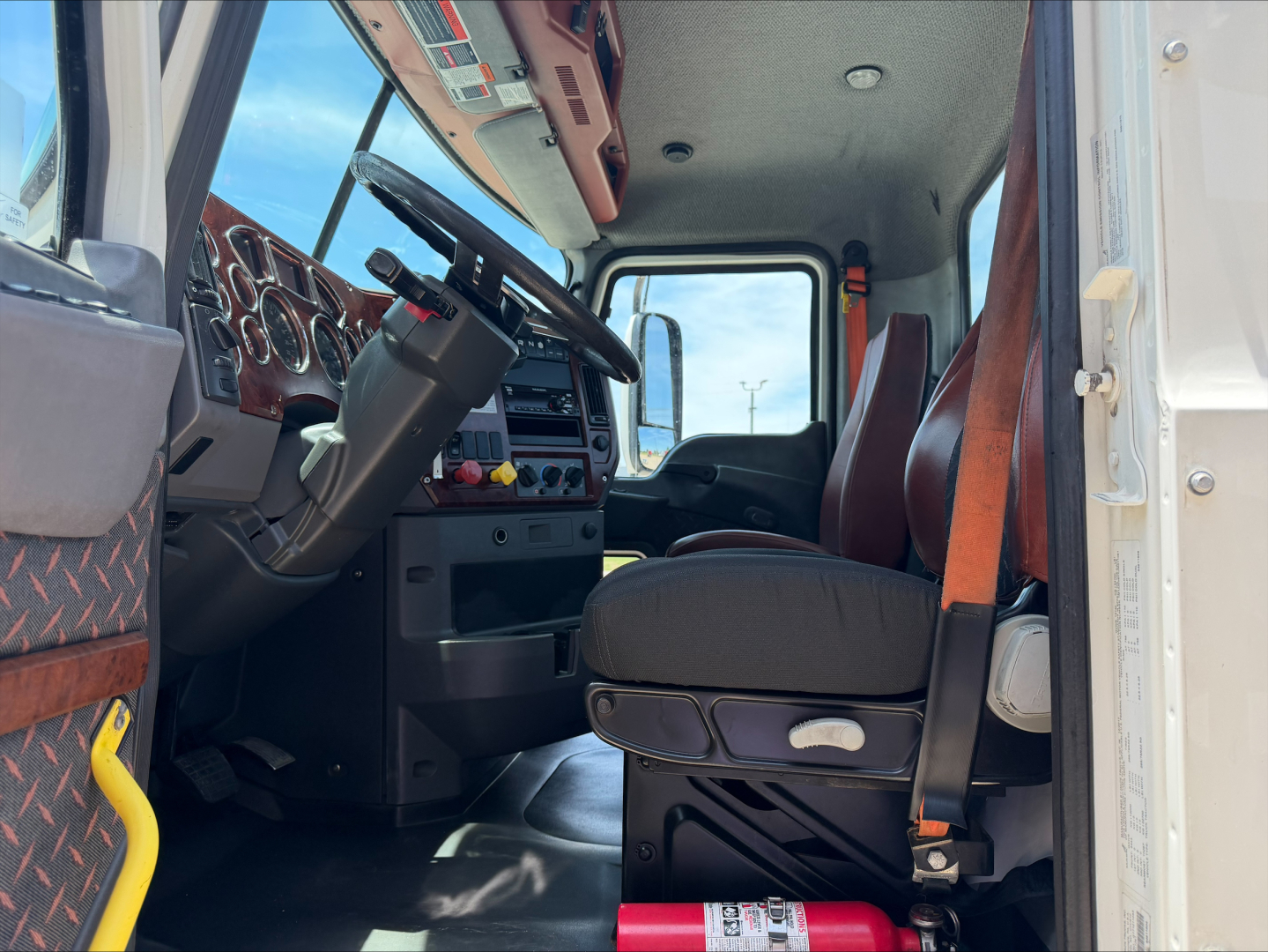 Truck cab interior of a 2018 Mack CXU612 showing driver seat, steering wheel, dashboard and door; spacious cab with brown diamond-plate panels.