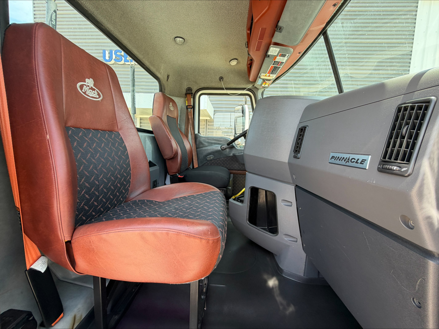 Truck cab interior showing driver's seat and passenger seat with red upholstery; dashboard with Pinnacle badge on glove compartment; overhead console and window; general cab interior of a Mack CXU612.
