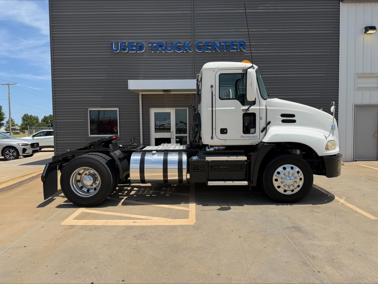 2018 Mack CXU612 daycab truck side view in front of a used truck center building; visible text includes USED TRUCK CENTER on the building sign; chrome fuel tank and wheels visible.