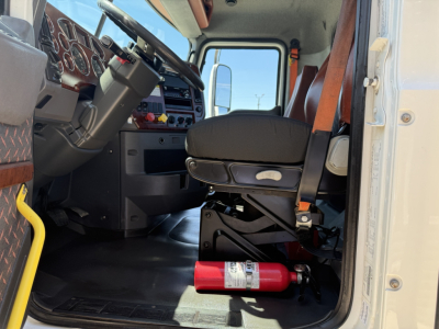 Truck cab interior of a 2018 Mack CXU612 from driver side view with steering wheel, dashboard, and a red fire extinguisher under the seat.