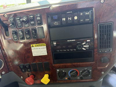 Truck dash panel showing multiple switches, a Mack stereo, climate controls, and warning label on woodgrain dashboard; gear indicator shows N and R with plus/minus controls.