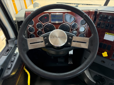 Truck cab interior with steering wheel and dashboard gauges; central steering wheel has 'MACK' logo. Dashboard features multiple round gauges, wood-grain panel, and various switches; windshield and passenger area visible.